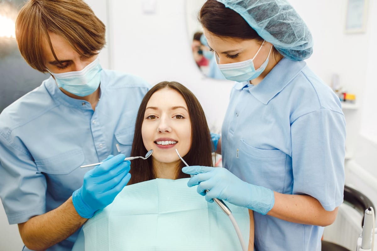 A dentist and an assistant examining a seated patient's teeth with a mirror and probe
